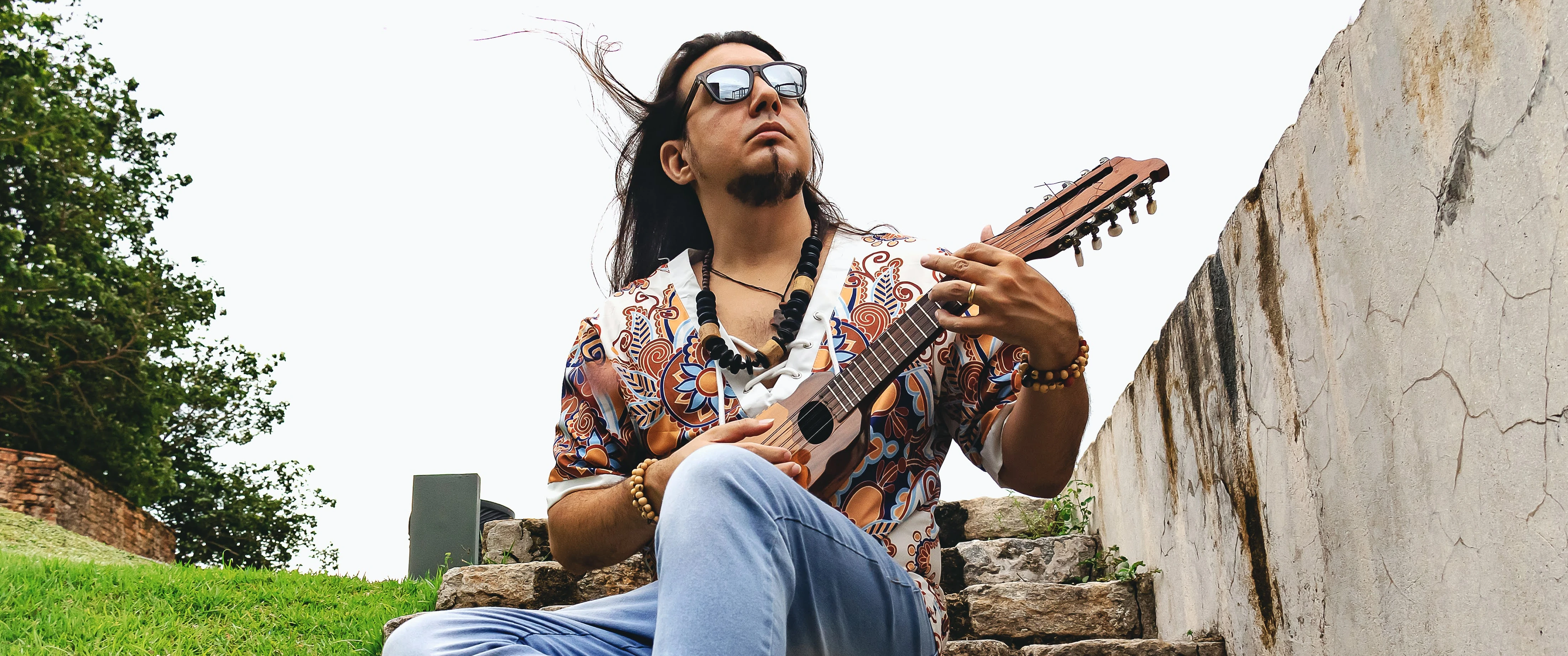 O artista sentado está sentado numa escadaria de pedra, de óculos escuros, camisa estampada e tocando um instrumento de cordas.