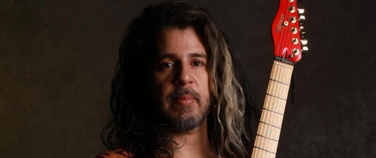 O músico está de cabelo solto e guitarra vermelha nas mãos.