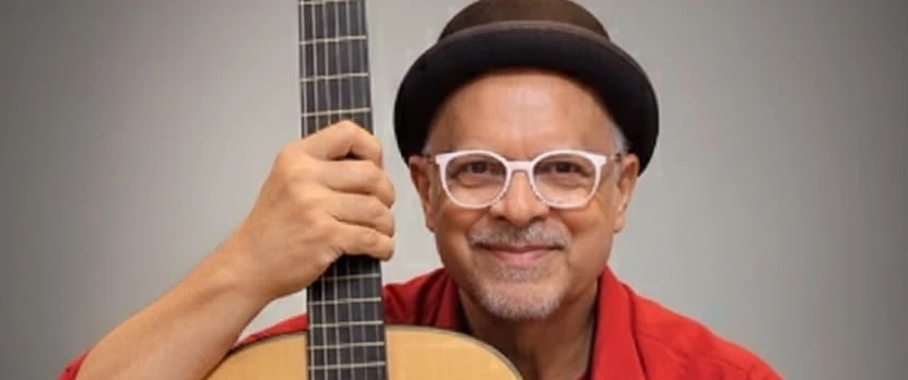 O músico está com camisa vermelha, de óculos branco, sorrindo e segurando o seu violão.