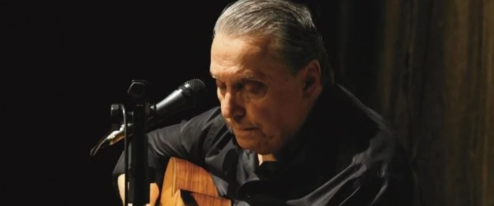 O músico está com camisa escura, na contra-luz, tocando violão.