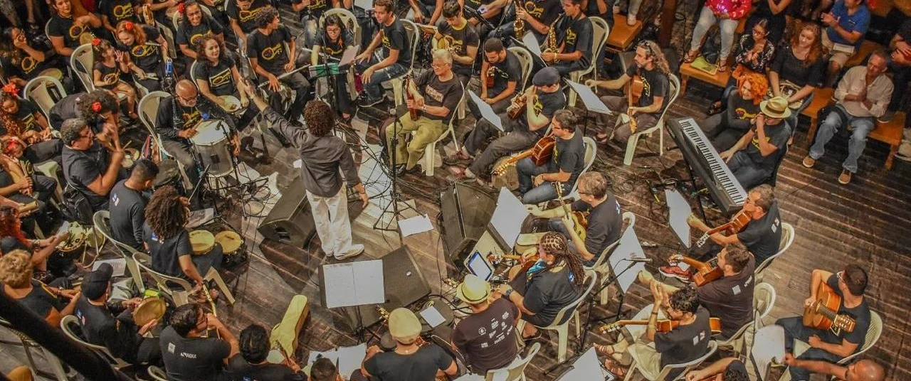 Grupo reunido em formato de Roda, com regente ao meio de camisa preta e calça branca. 